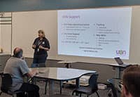 A person stands at the front of a classroom giving a presentation at the UEN Tech Summit. Several people sit at tables, and a large projected slide titled Utility Support is displayed behind the presenter.