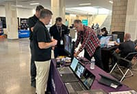 Four men stand around a table with laptops and promotional materials at the Tech Summit, engaging in conversation and demonstrations. Other booths and attendees are visible in the background at this dynamic UEN event.