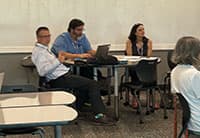 Three people sit at a table with laptops and papers in a classroom, attending the UEN Tech Summit. Another person is partially visible in the foreground as the group listens to a discussion.