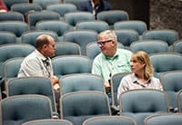 Three people sit and talk in a mostly empty auditorium with blue seats at the Tech Summit; two men face each other while a woman sits beside them.