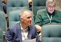 At the UEN Tech Summit, two men sit in auditorium chairs; one in front with a blue blazer holding papers and glancing to the side, while another behind him in a green shirt looks down at an electronic device.