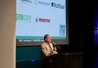 A person in a tan suit stands at a podium, speaking into a microphone at the Tech Summit, with a projection screen behind displaying several company logos and the UEN branding.