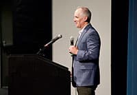A man in a blue blazer stands at a podium, smiling and holding a microphone as he speaks on stage at the UEN Tech Summit in front of a blank screen.