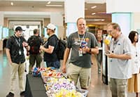A group of people stands around a snack table at the UEN Tech Summit; some are eating, talking, and wearing lanyards with UEN logos, with snacks displayed in bowls on the table.
