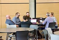 Six people sit around a table in a modern, open area at the UEN Tech Summit, engaged in lively conversation. Some have laptops open in front of them against light-colored panel walls in the background.