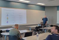 A man stands at the front of a modern classroom at the UEN Tech Summit, presenting to a few seated attendees. A large screen displays a diagram and text, while white walls and several tables and chairs complete the setting.