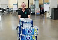 A smiling person in a black shirt pushes a cart stacked with several cases of bottled water through a spacious indoor area at the annual Tech Summit, with tables and chairs in the background, organized by UEN.