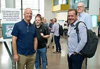 A group of four men stand indoors at the UEN Tech Summit in a bright, modern building, smiling at the camera. Other people and informational signs are visible in the background.