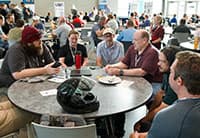 Six people sit around a table in a busy, bright cafeteria at the Tech Summit, engaged in conversation and smiling. Plates and drinks are on the table, with other UEN groups visible in the background.