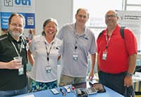 Four adults stand smiling behind a table with tech gadgets and name badges at the Tech Summit. A display and UTN informational materials are visible in the background.