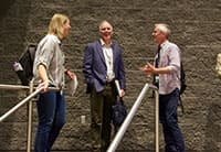 Three people stand and talk on a staircase in front of a textured wall at the Tech Summit, two men and one woman, all wearing UEN conference badges and carrying bags.