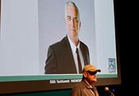 At the Tech Summit, a man in a cap stands at a podium before a large screen displaying a professional portrait of another man in a suit and tie.