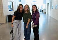 Three women stand together smiling in a brightly lit hallway at the Tech Summit. One holds a camera, and they are casually dressed. The background features open doors and some hallway fixtures.