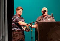 Two men stand on stage at the UEN Tech Summit shaking hands as one receives an award. Both wear lanyards; one wears a striped shirt, and the other a sports hat and polo. A microphone and podium are in front of them, with a teal background.