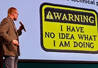 At the UEN Tech Summit, a person holding a microphone stands next to a presentation slide displaying a Warning sign that reads: I have no idea what I am doing.