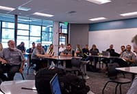 A group of people sit at round tables in a classroom or meeting room during the UEN Tech Summit, attentively listening to someone out of view. Large windows let in natural light and a whiteboard is visible on the wall.