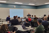 At the UEN Tech Summit, a speaker presents information on a screen to a classroom of attentive adults seated at tables with laptops and notebooks.