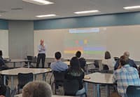 A man stands and gestures while giving a presentation at the UEN Tech Summit to a group of people seated at tables in a classroom. A colorful slide is projected on a whiteboard behind him.