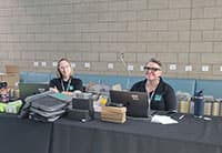 Two people sit behind a table covered with laptops, paperwork, and supplies at the UEN Tech Summit, smiling at the camera. Behind them is a wall with electrical outlets and a stack of boxes.