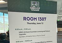 A sign displays the schedule for Room 1307 at the UEN Tech Summit, listing sessions and times for Thursday, June 12, including a Zoom Essentials session and speakers’ names, with windows and people in the background.