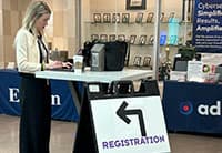 A woman stands at a high table with a laptop and coffee cup. In front of the table is a sign with a left arrow and the word Registration, guiding attendees at the Tech Summit. Display booths are visible in the background.