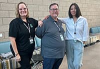 Three people stand together indoors at the UEN Tech Summit, smiling at the camera. Two wear dark shirts and one wears a white blouse and light pants, posing in front of a brick wall with lounge chairs in the background.