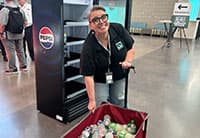 A smiling woman wearing glasses and a black shirt stands next to an empty Pepsi cooler, holding a red cart filled with bottles and cans at the UEN Tech Summit in a spacious indoor setting.